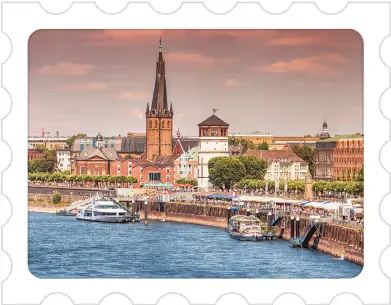 Riverfront cityscape with boats docked along the quay and a tall church spire under a cloudy sky.