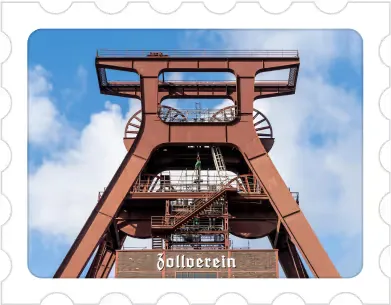 Rust-colored industrial mining tower structure against a blue sky with clouds, labeled 'Zollverein'.