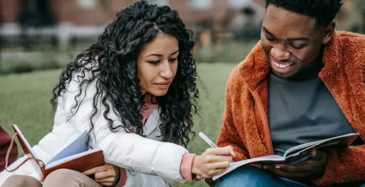 Two young adults outdoors studying together with open notebooks, one pointing at the book as they discuss.